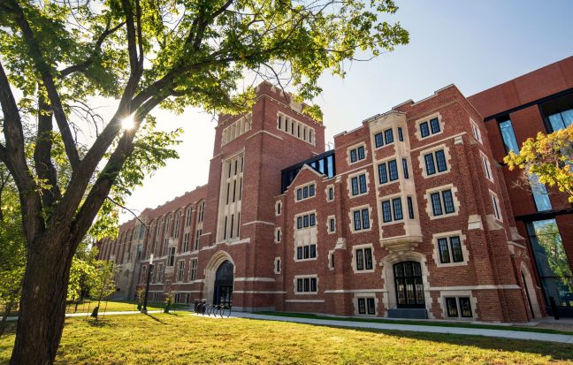 A 100 year old building in red bricks with white accents featuring many windows, a central tower, and new, modern additions. There is a tree in the foreground and the sky is clear.